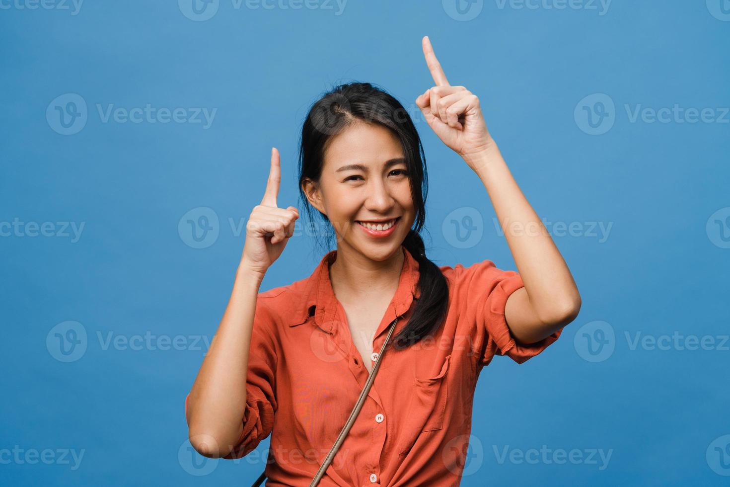 Portrait of young Asian lady smiling with cheerful expression, shows something amazing at blank space in casual cloth and looking at camera isolated over blue background. Facial expression concept. photo