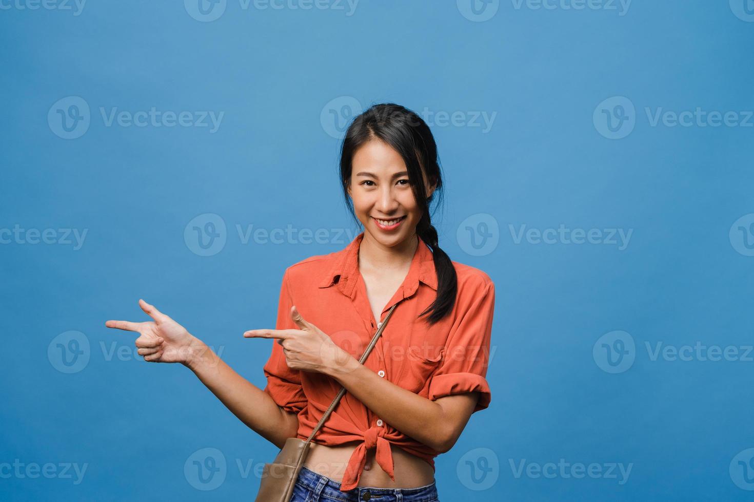 Portrait of young Asian lady smiling with cheerful expression, shows something amazing at blank space in casual cloth and looking at camera isolated over blue background. Facial expression concept. photo