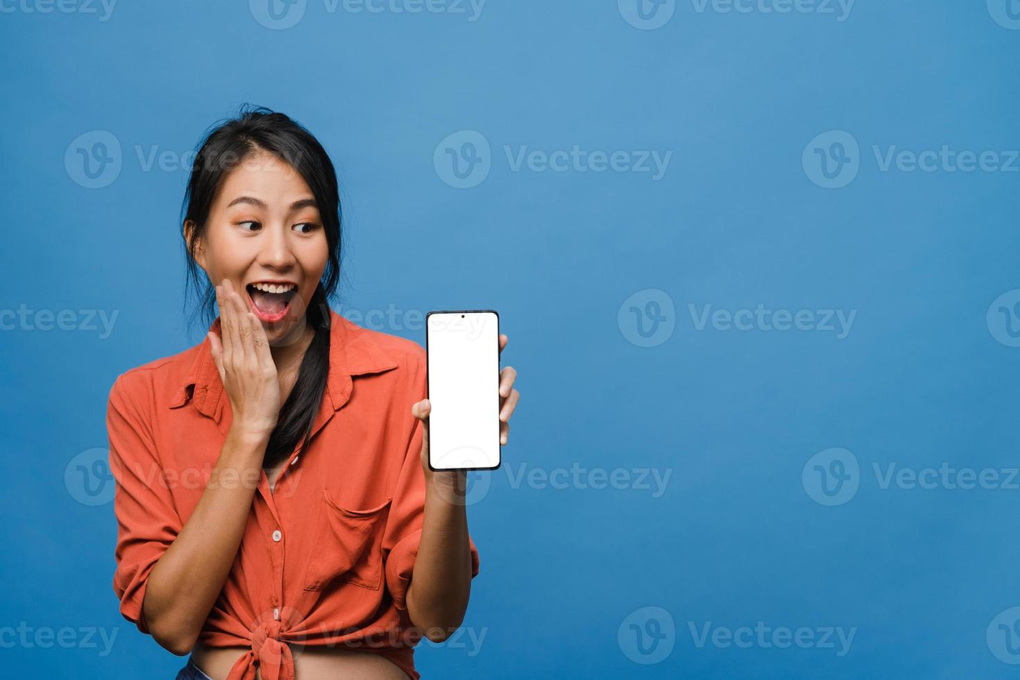 Joven asiática muestra la pantalla vacía del teléfono inteligente con expresión positiva, sonríe ampliamente, vestida con ropa casual sintiendo felicidad sobre fondo azul. teléfono móvil con pantalla en blanco en mano femenina. foto