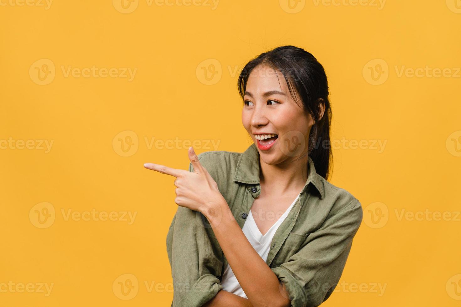 Portrait of young Asian lady smiling with cheerful expression, shows something amazing at blank space in casual clothing and standing isolated over yellow background. Facial expression concept. photo