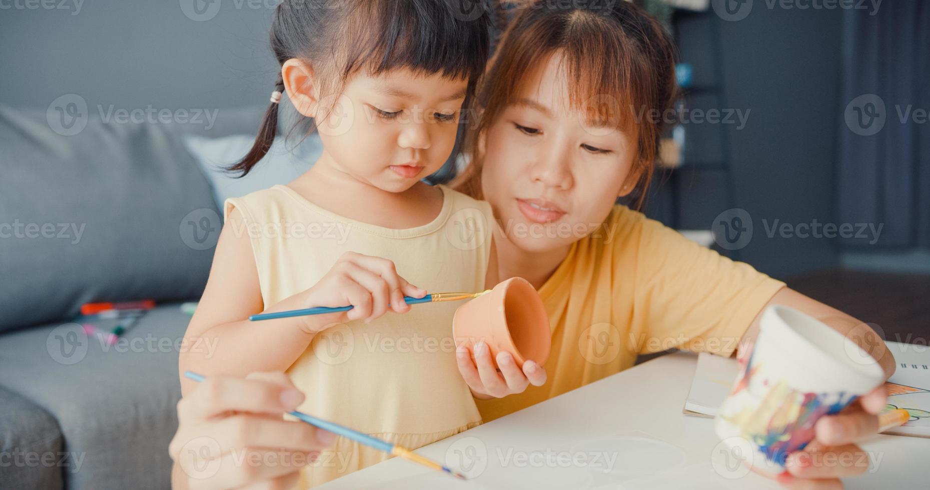 Happy cheerful Asia family mom teach toddler girl paint ceramic pot having fun relax on table in living room at house. Spending time together, Social distance, Quarantine for coronavirus prevention. photo