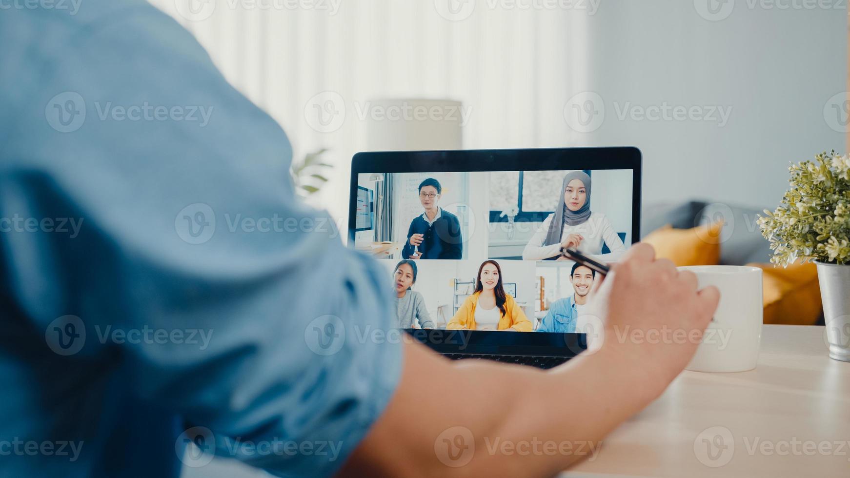 joven empresario de asia usando laptop hablar con colegas sobre el plan en la reunión de videollamada mientras trabaja desde casa en la sala de estar. autoaislamiento, distanciamiento social, cuarentena para la prevención del coronavirus. foto