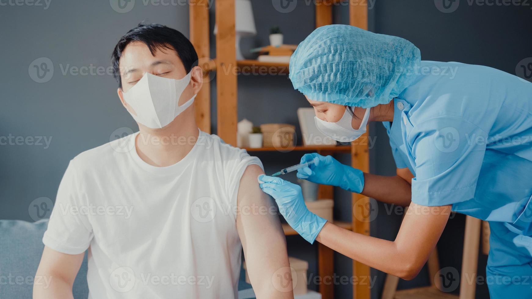 Young Asia lady nurse giving Covid-19 or flu antivirus vaccine shot to male patient wear face mask protection from virus disease sit on couch in living room at house. Vaccination concept. photo