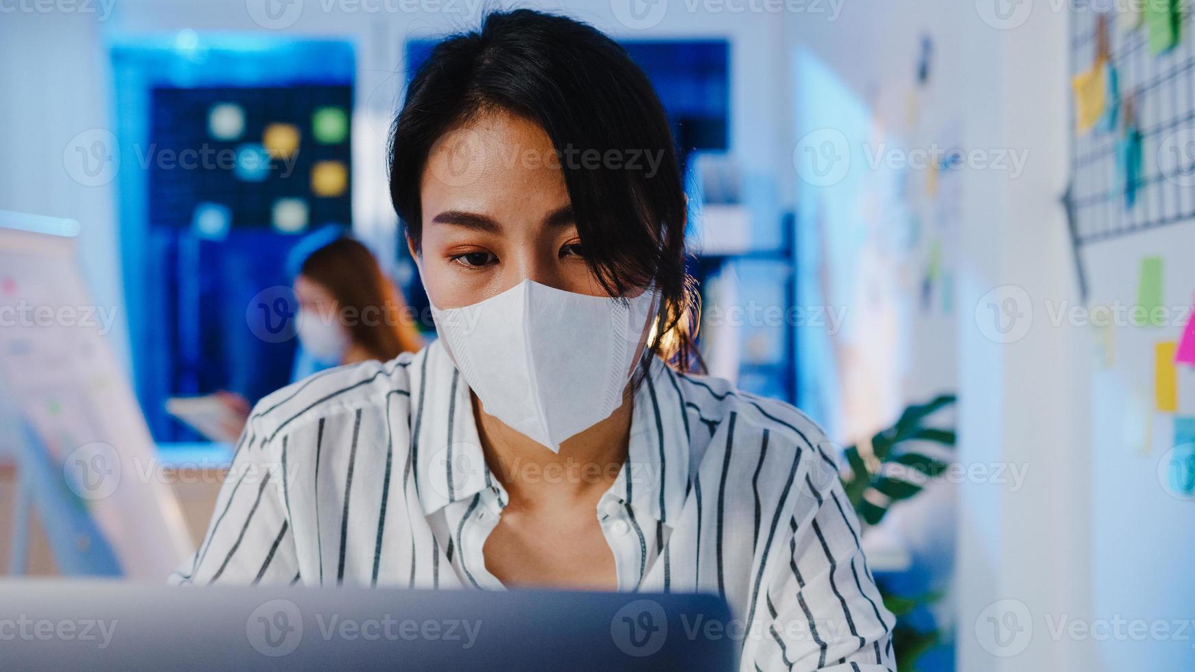 Happy Asia businesswoman wearing medical face mask for social distancing in new normal situation for virus prevention while using laptop back at work in office night. Life and work after coronavirus. photo