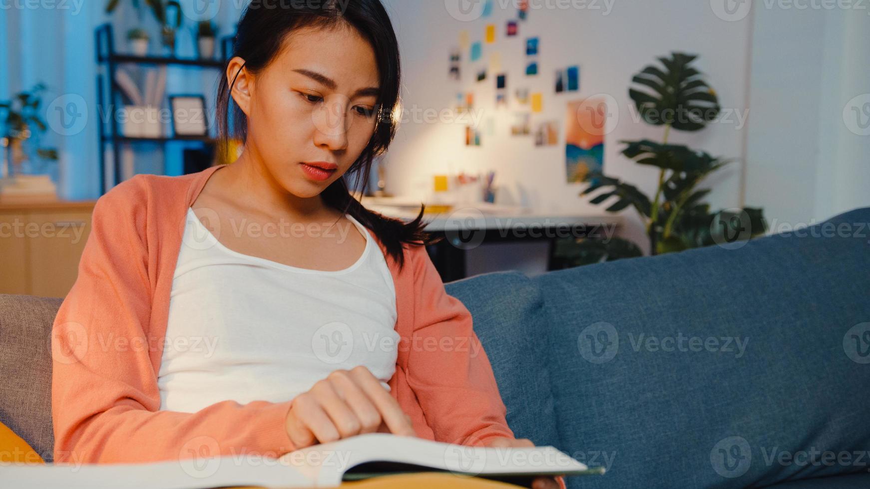 Night time beautiful Asia lady read book with happiness in peaceful living room on sofa. Education from home, Stay at house, Self quarantine activity, Fun activity for covid or coronavirus quarantine. photo