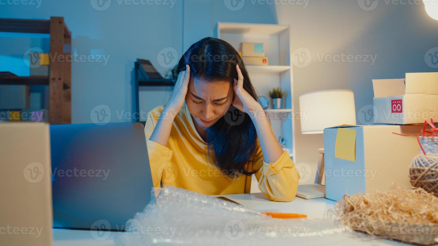 Young Asia businesswoman look around room full of product stuff and parcel box feel stress and upset with bad sell in home office at night. Small business owner, online market delivery concept. photo