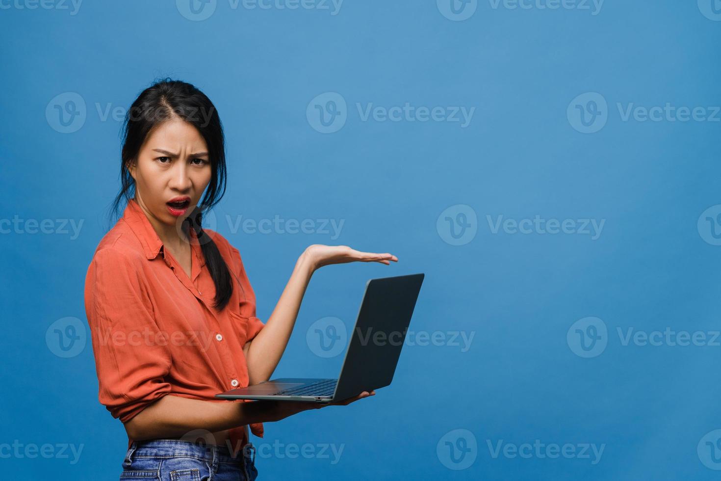 Jovencita asiática usando laptop con expresión negativa, gritando emocionado, llorando emocionalmente enojado en ropa casual y parado aislado sobre fondo azul con espacio de copia en blanco. concepto de expresión facial. foto