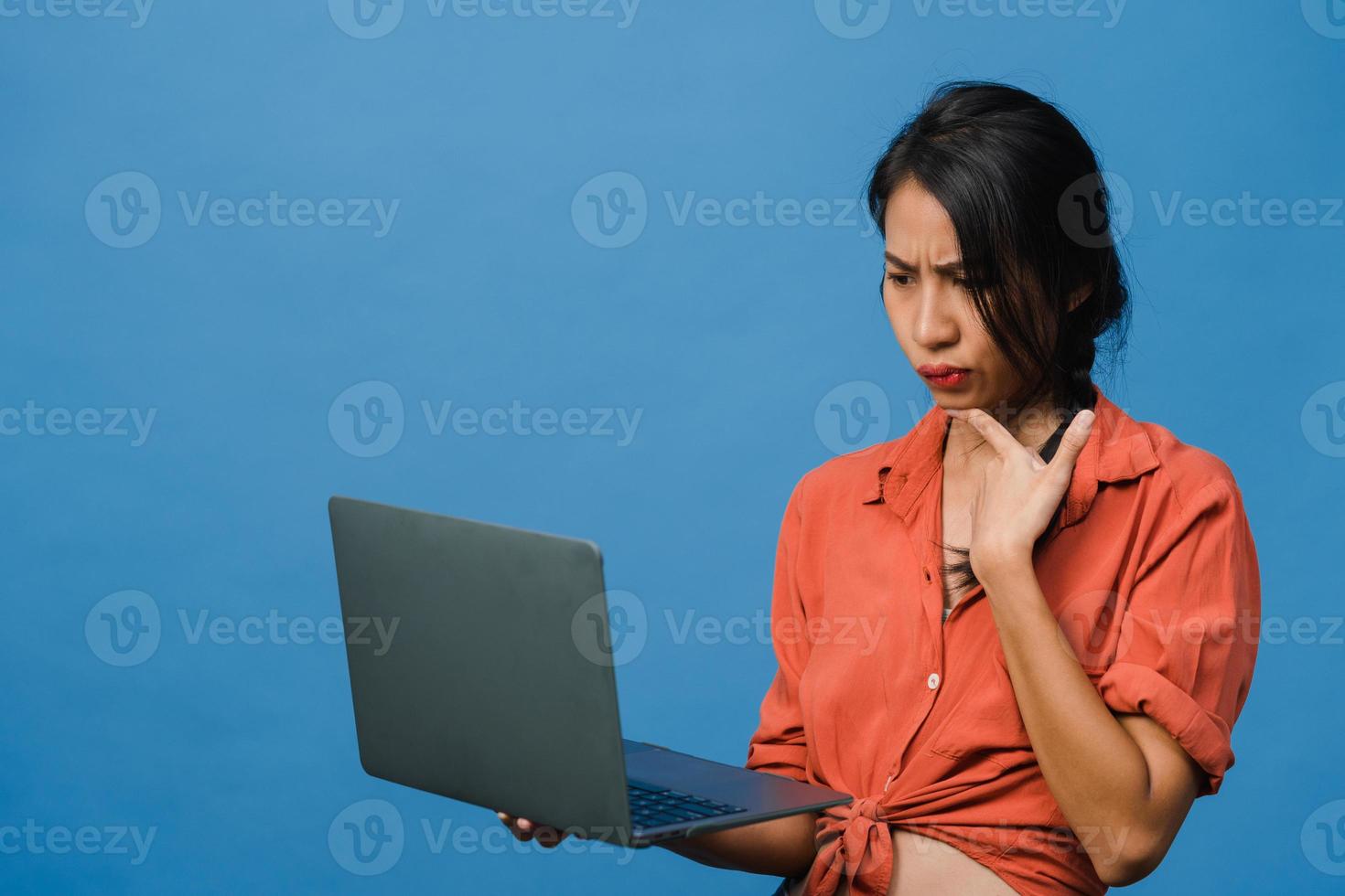 Jovencita asiática usando laptop con expresión negativa, gritando emocionado, llorando emocionalmente enojado en ropa casual y parado aislado sobre fondo azul con espacio de copia en blanco. concepto de expresión facial. foto