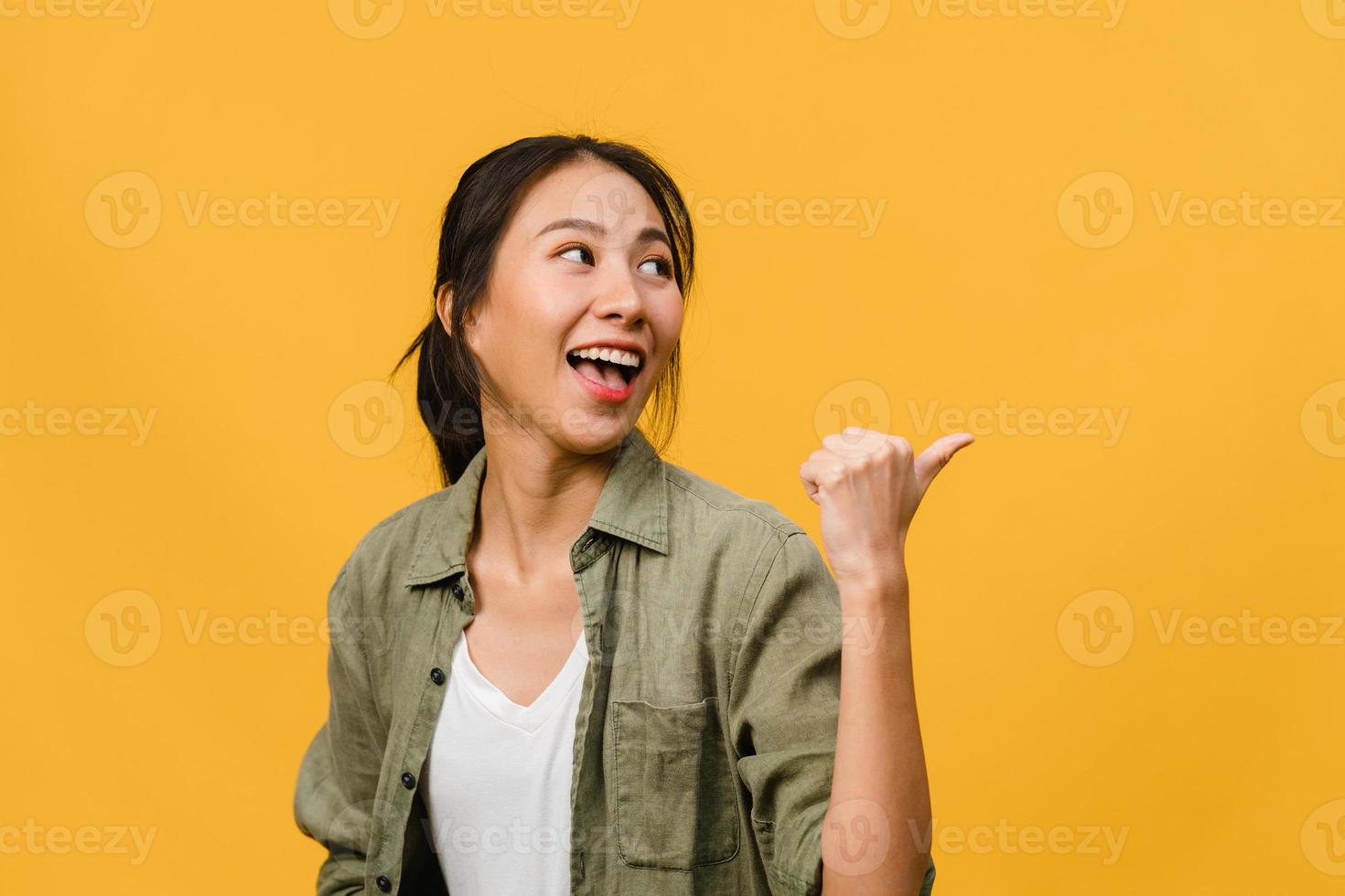 Portrait of young Asian lady smiling with cheerful expression, shows something amazing at blank space in casual clothing and standing isolated over yellow background. Facial expression concept. photo