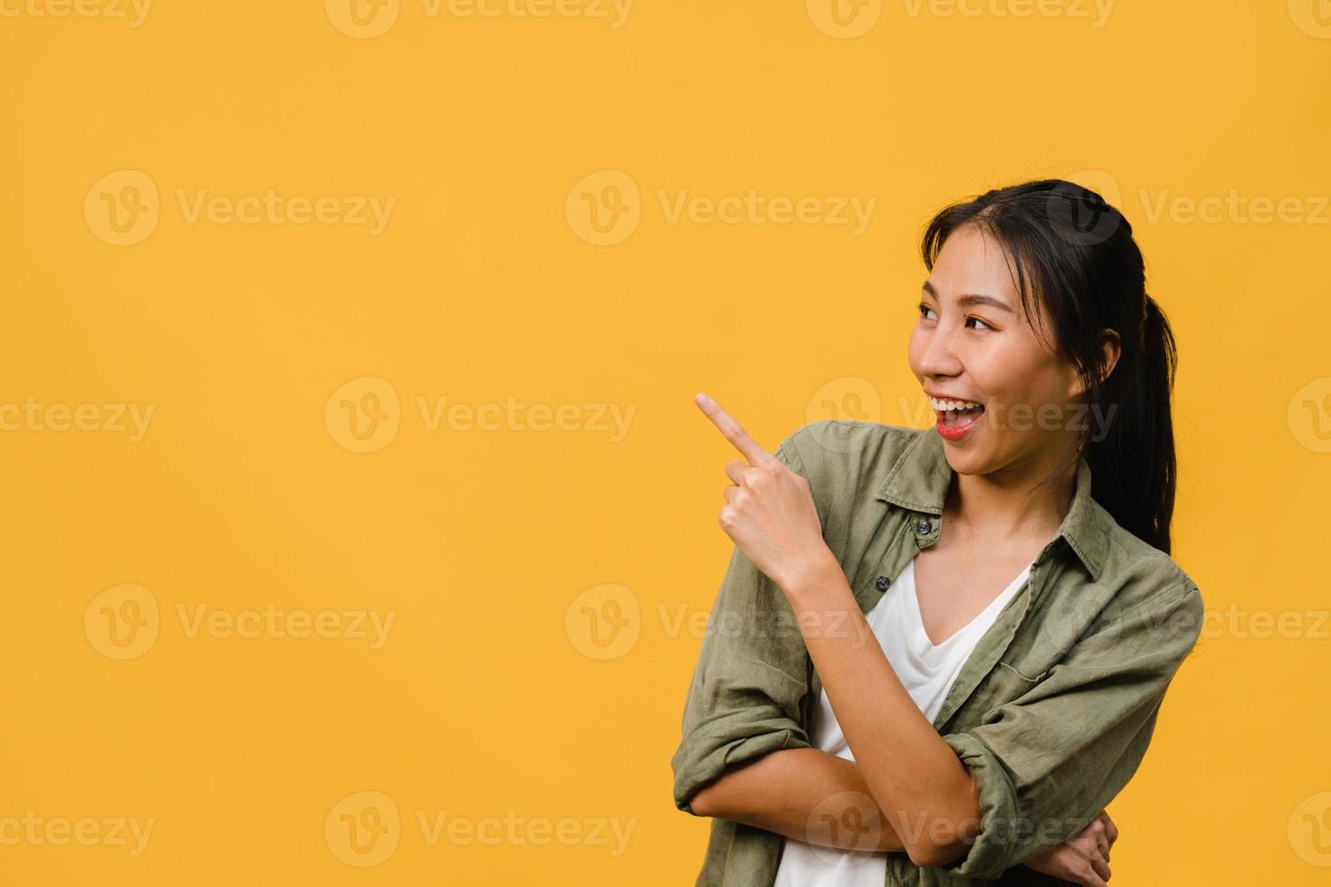 Portrait of young Asian lady smiling with cheerful expression, shows something amazing at blank space in casual clothing and standing isolated over yellow background. Facial expression concept. photo