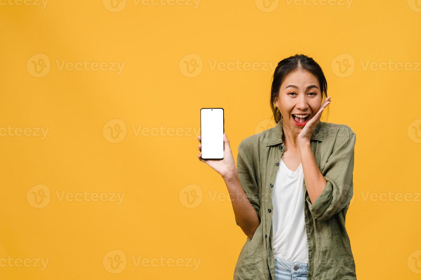 Joven asiática muestra la pantalla vacía del teléfono inteligente con expresión positiva, sonríe ampliamente, vestida con ropa casual sintiendo felicidad sobre fondo amarillo. teléfono móvil con pantalla en blanco en mano femenina. foto