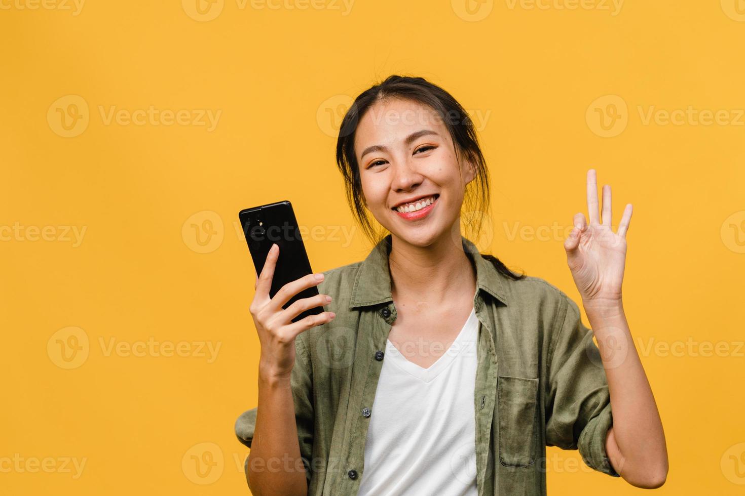 Sorprendió a la joven asiática que usa el teléfono móvil con expresión positiva, sonríe ampliamente, vestida con ropa casual y mirando a la cámara sobre fondo amarillo. feliz adorable mujer alegre se regocija con el éxito. foto