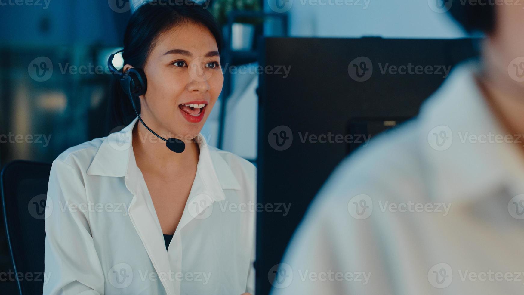 Millennial Asia young call center team or customer support service executive using computer and microphone headset working technical support in late night office. Telemarketing or sales job concept. photo