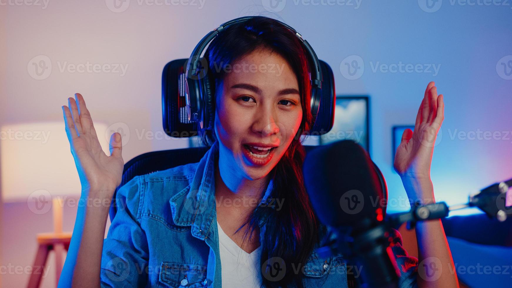 Happy asia girl blogger music influencer looking at camera broadcast record wear headphone online live talk in microphone with audience in living room home studio at night. Content creator concept. photo