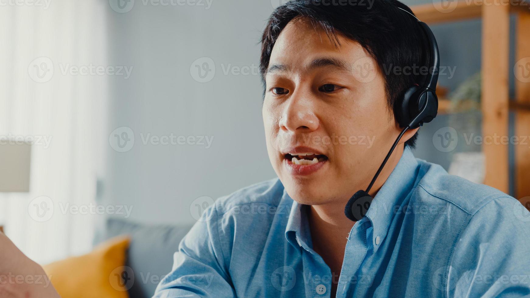 Young Asia businessman wear headphones using laptop talk to colleagues about plan in video call while work from home at living room. Self-isolation, social distancing, quarantine for covid prevention. photo