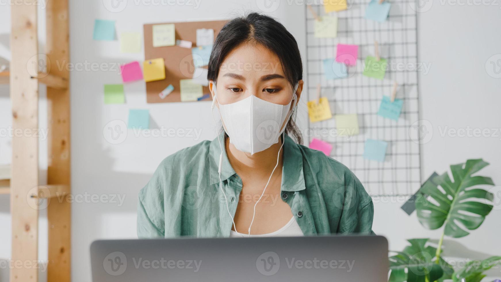 Asia businesswoman wearing medical face mask using laptop talk to colleagues about plan in video call while working from home at living room. Social distancing, quarantine for corona virus prevention. photo