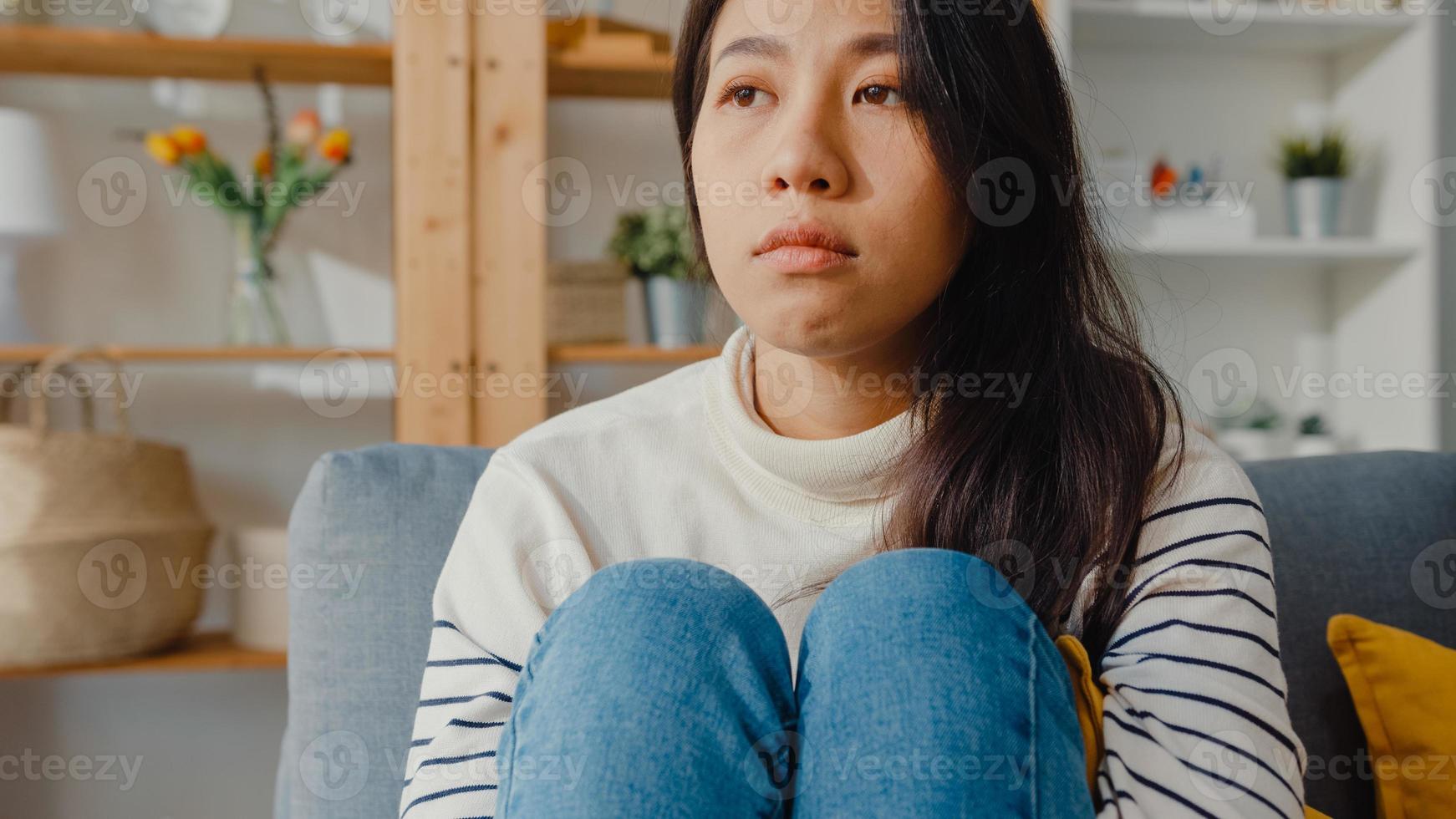 Thoughtful Asia lady sit embracing knees at sofa in living room at house look outside with feel lonely, Sad depressed teenager spend time alone stay at home, Social distance, Coronavirus Quarantine. photo