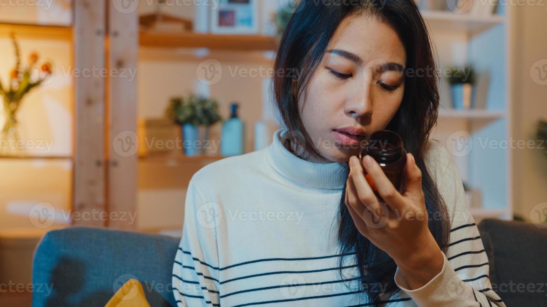 Sick asian young lady holding pill take a look medicine sit on couch at home night. Girl taking medicine after doctor order, quarantine at home, Coronavirus social distancing healthcare concept. photo