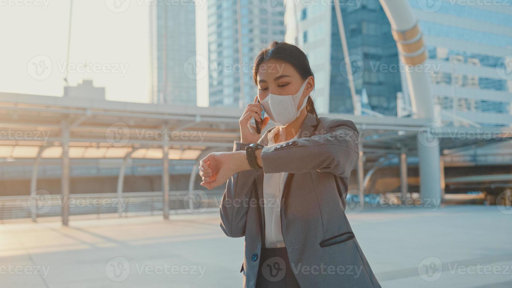 Young Asia businesswoman in fashion office clothe wear medical face mask talk via phone while walk alone outdoor in urban city. Business on go, Social distancing to prevent spread of COVID-19 concept. photo