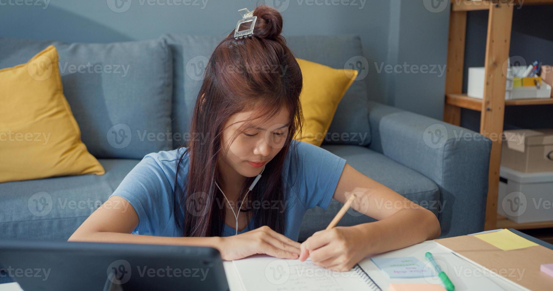 Young Asia girl teenager with casual wear headphones use digital tablet learn online write lecture notebook in living room at house. Isolate education online e-learning coronavirus pandemic concept. photo
