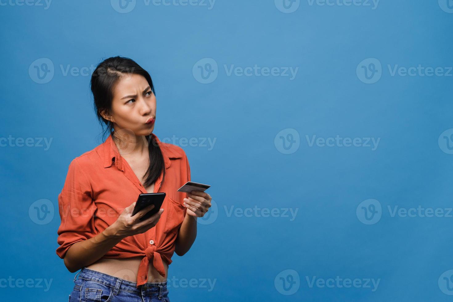 Jovencita asiática usando teléfono y tarjeta bancaria de crédito con expresión negativa, gritos emocionados, gritos emocionales enojados en ropa casual y de pie aislado sobre fondo azul. concepto de expresión facial. foto