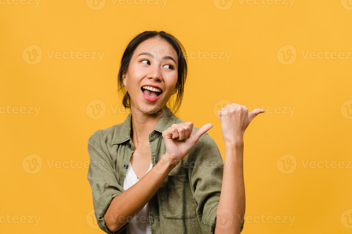 Portrait of young Asian lady smiling with cheerful expression, shows something amazing at blank space in casual clothing and standing isolated over yellow background. Facial expression concept. photo