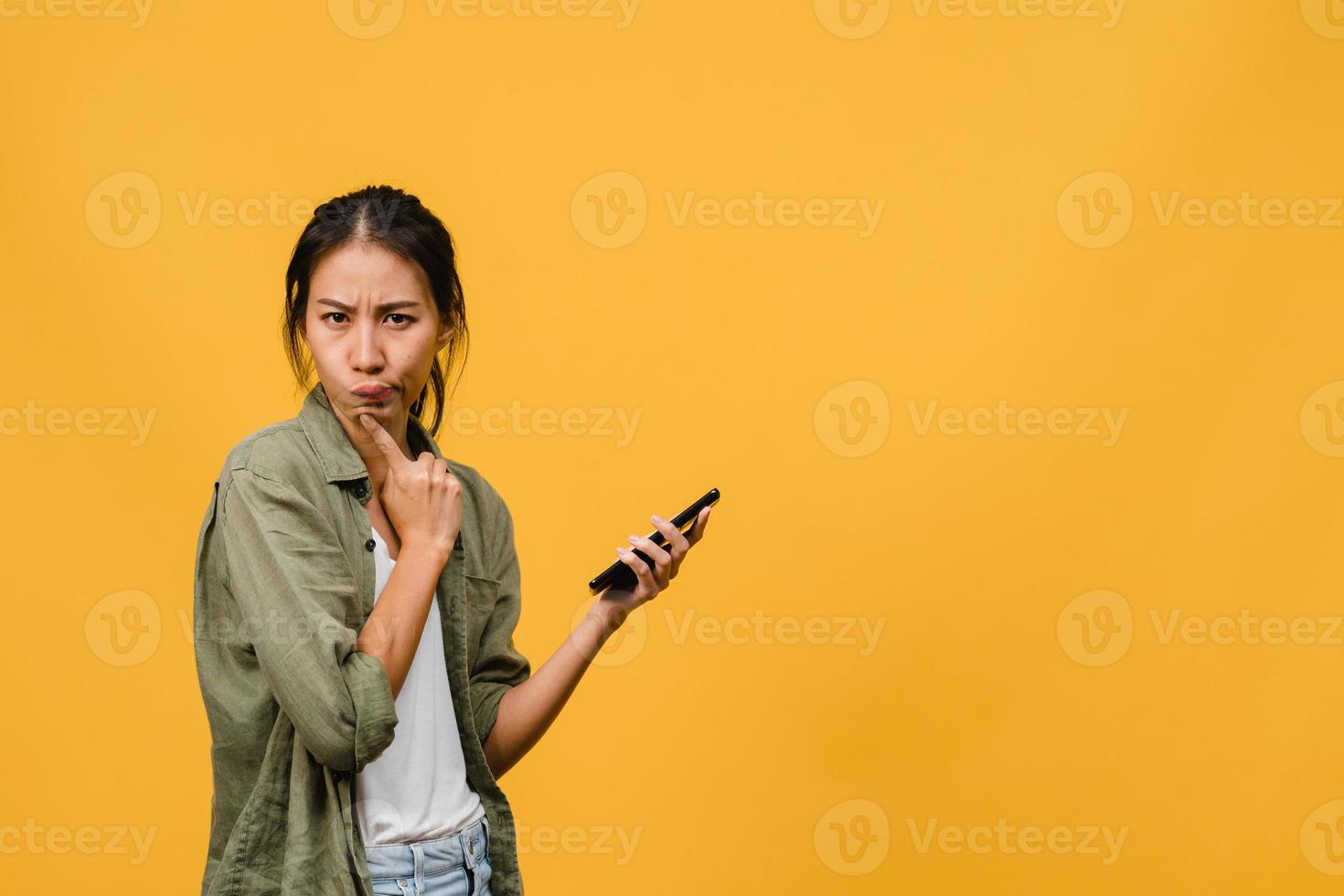 Thinking dreaming young Asia lady using phone with positive expression, dressed in casual cloth feeling happiness and stand isolated on yellow background. Happy adorable glad woman rejoices success. photo