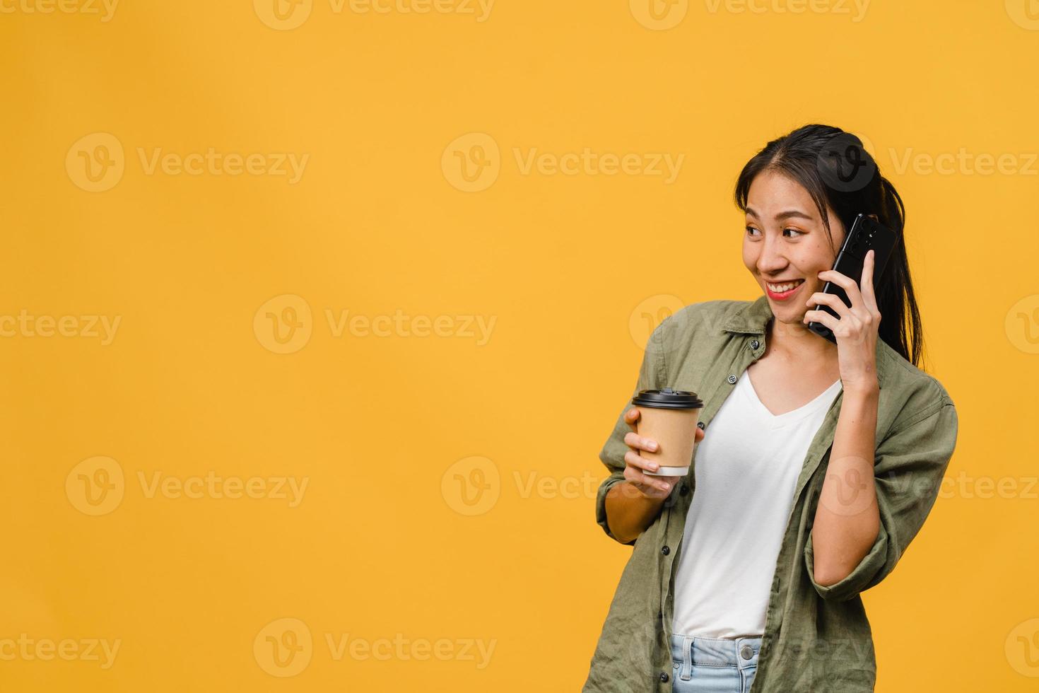 La joven asiática habla por teléfono y sostiene la taza de café con expresión positiva, sonríe ampliamente, vestida con un paño informal sintiendo felicidad y parada aislada sobre fondo amarillo. concepto de expresión facial. foto