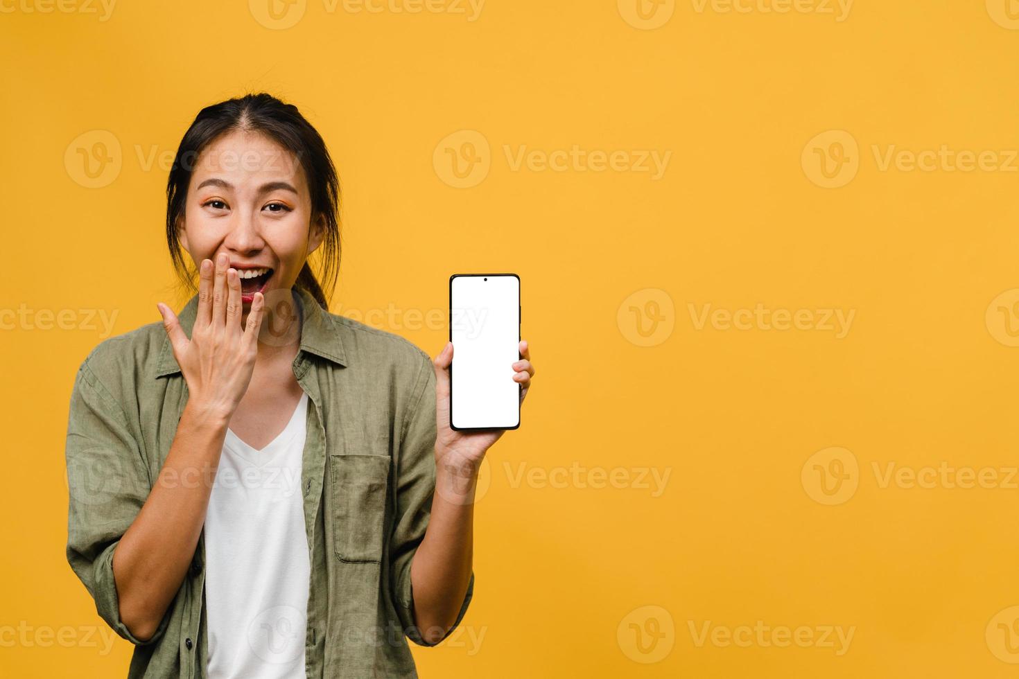 Young Asia lady show empty smartphone screen with positive expression, smiles broadly, dressed in casual clothing feeling happiness on yellow background. Mobile phone with white screen in female hand. photo