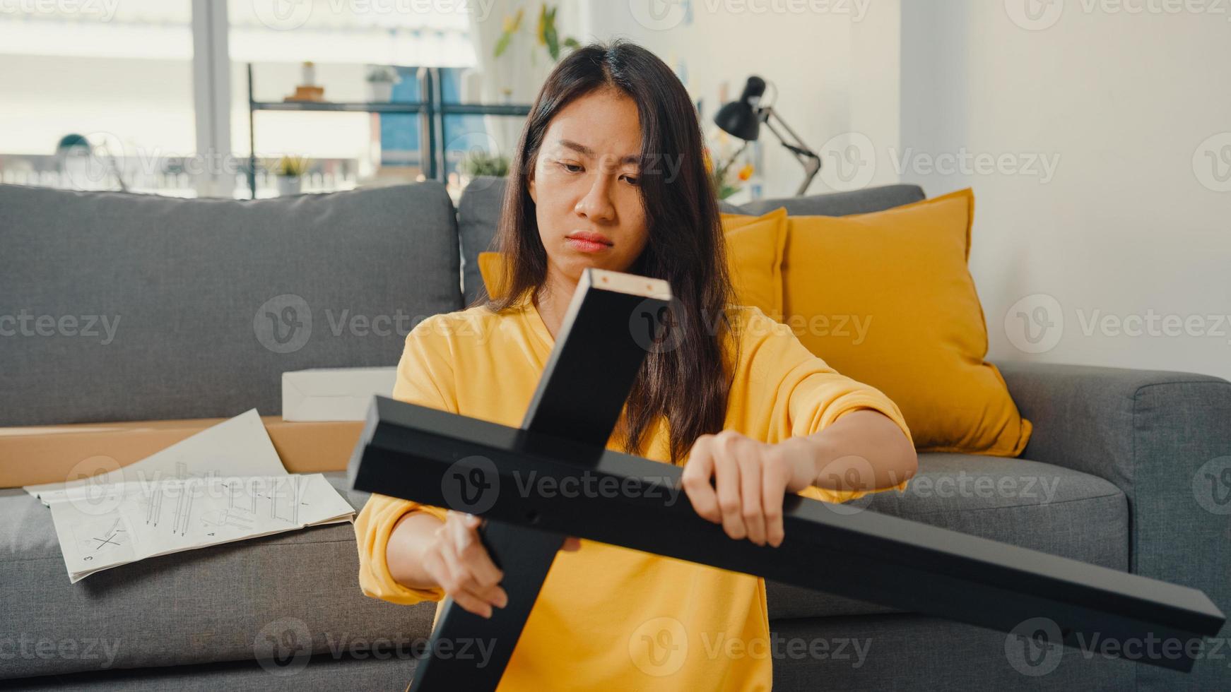 Feliz joven asiática desempacando la caja y leyendo las instrucciones para ensamblar muebles nuevos decorar la mesa de construcción de la casa con caja de cartón en la sala de estar de casa. foto