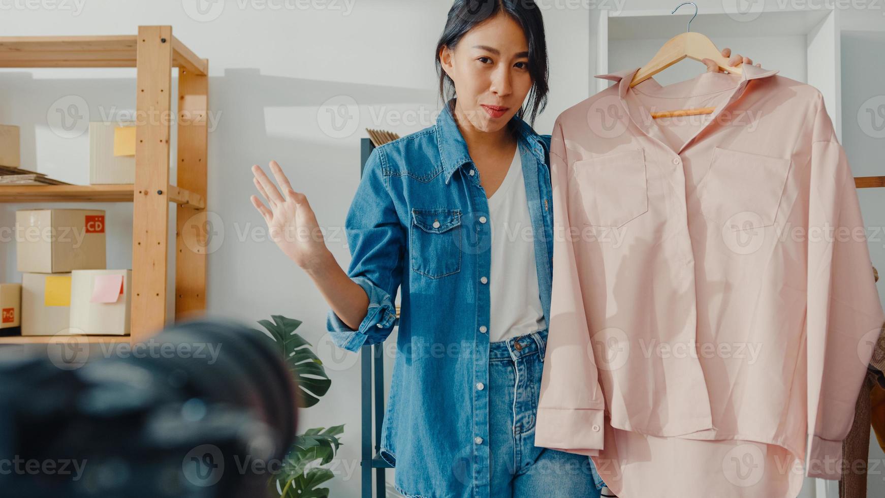 joven diseñadora de moda asiática que usa un teléfono móvil y recibe una orden de compra y muestra la ropa, grabando video en vivo en línea con la cámara. propietario de una pequeña empresa, concepto de entrega de mercado en línea. foto