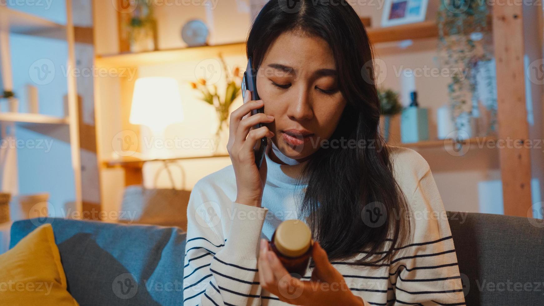 mujer asiática joven enferma mantenga la medicina sentarse en el sofá use el teléfono inteligente para consultar con el médico en la noche en casa. chica toma medicamentos después de la orden del médico, cuarentena en casa, concepto de coronavirus de distancia social. foto