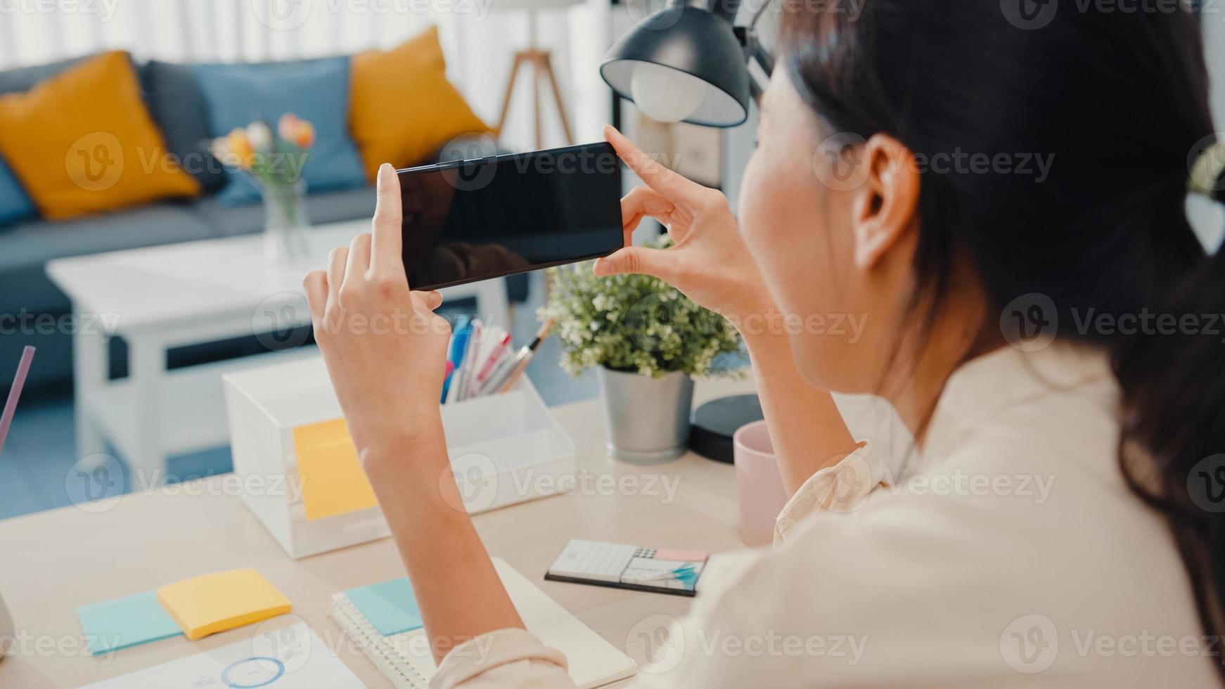 La jovencita asiática usa un teléfono inteligente con pantalla negra en blanco simulada para mostrar texto publicitario mientras trabaja de manera inteligente desde casa en la sala de estar. tecnología chroma key, concepto de diseño de marketing. foto