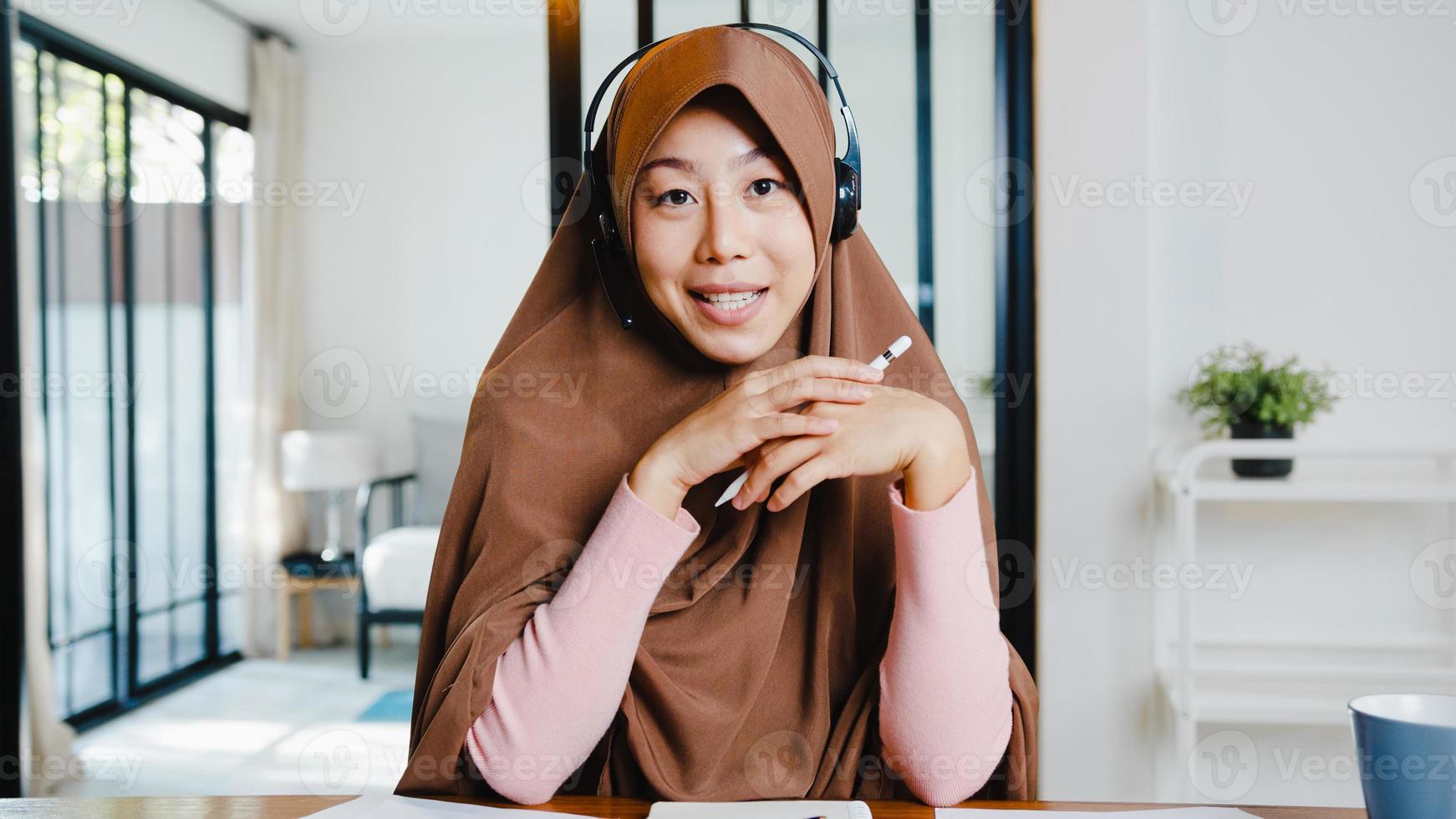 Asia muslim lady wear headphone using computer laptop talk to colleagues about sale report in video call while remotely work from home at living room. Social distancing, quarantine for corona virus. photo