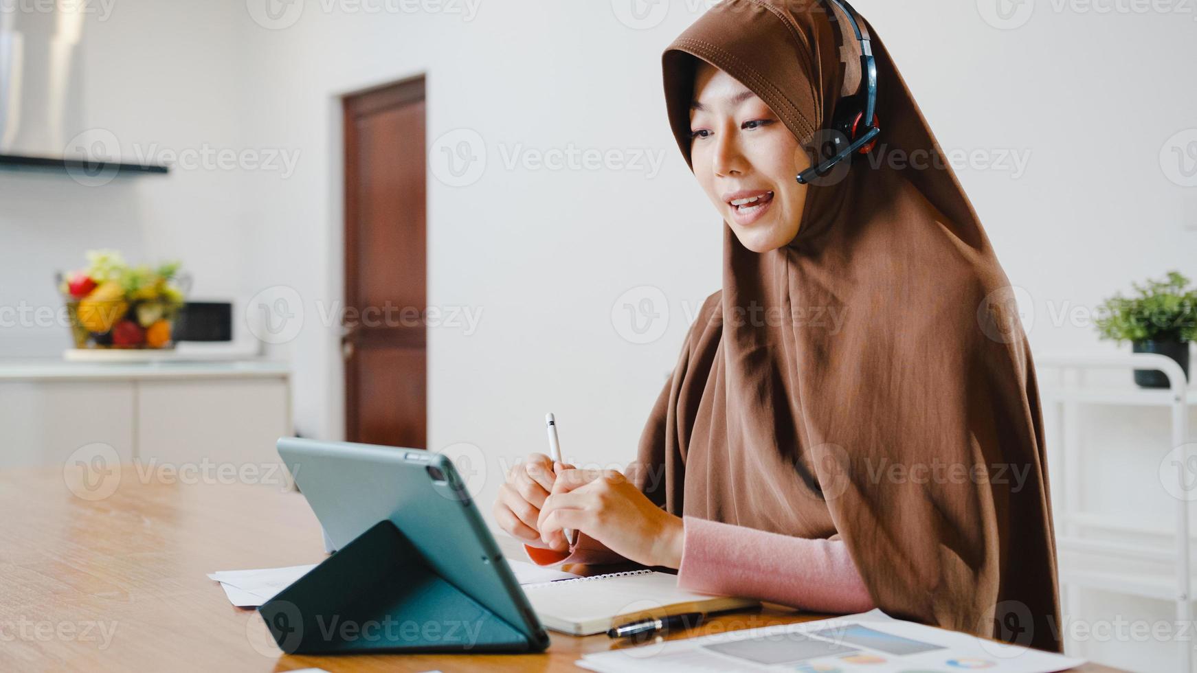 Asia muslim lady wear headphone using digital tablet talk to colleagues about sale report in conference video call while working from home at kitchen. Social distancing, quarantine for corona virus. photo