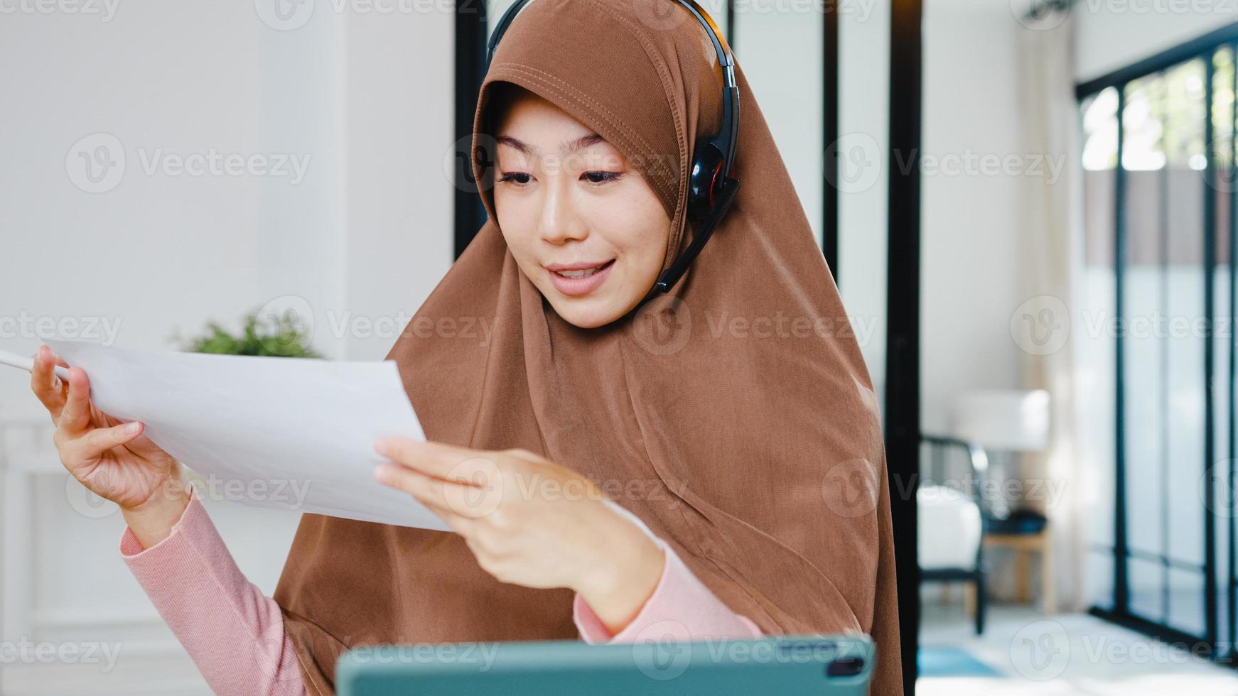 Asia dama musulmana usa auriculares con tableta digital, hable con sus colegas sobre el informe de venta en una videollamada de conferencia mientras trabaja desde casa en la cocina. distanciamiento social, cuarentena por coronavirus. foto