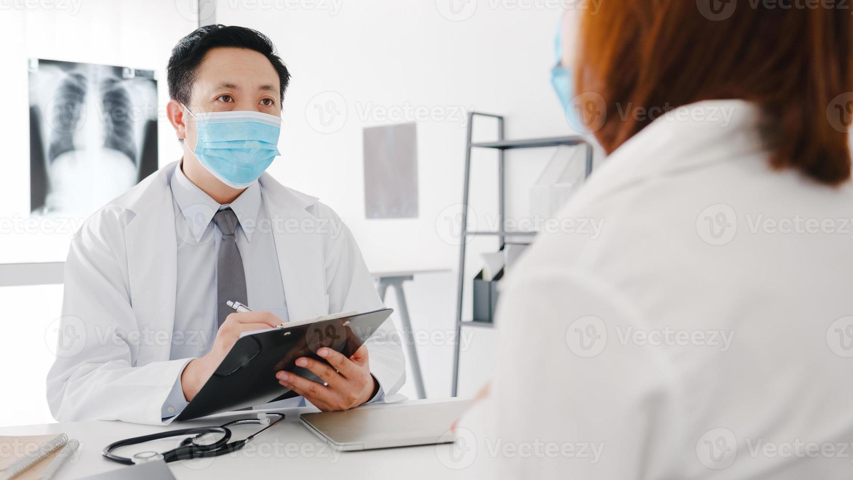 Serious Asia male doctor wear protective mask using clipboard is delivering great news talk discuss results or symptoms with female patient in hospital office. Lifestyle new normal after corona virus. photo