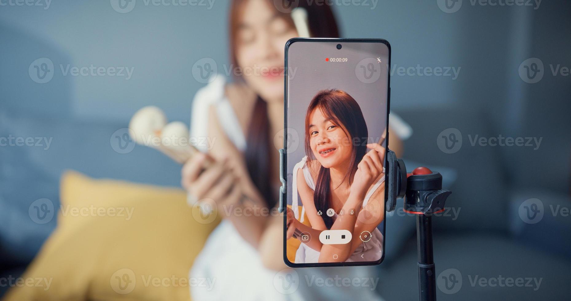 Happy young Asian girl makeup vlogger front of phone camera enjoy review talk with audience in living room at house. Social distance coronavirus pandemic concept. Freedom and active lifestyle concept. photo