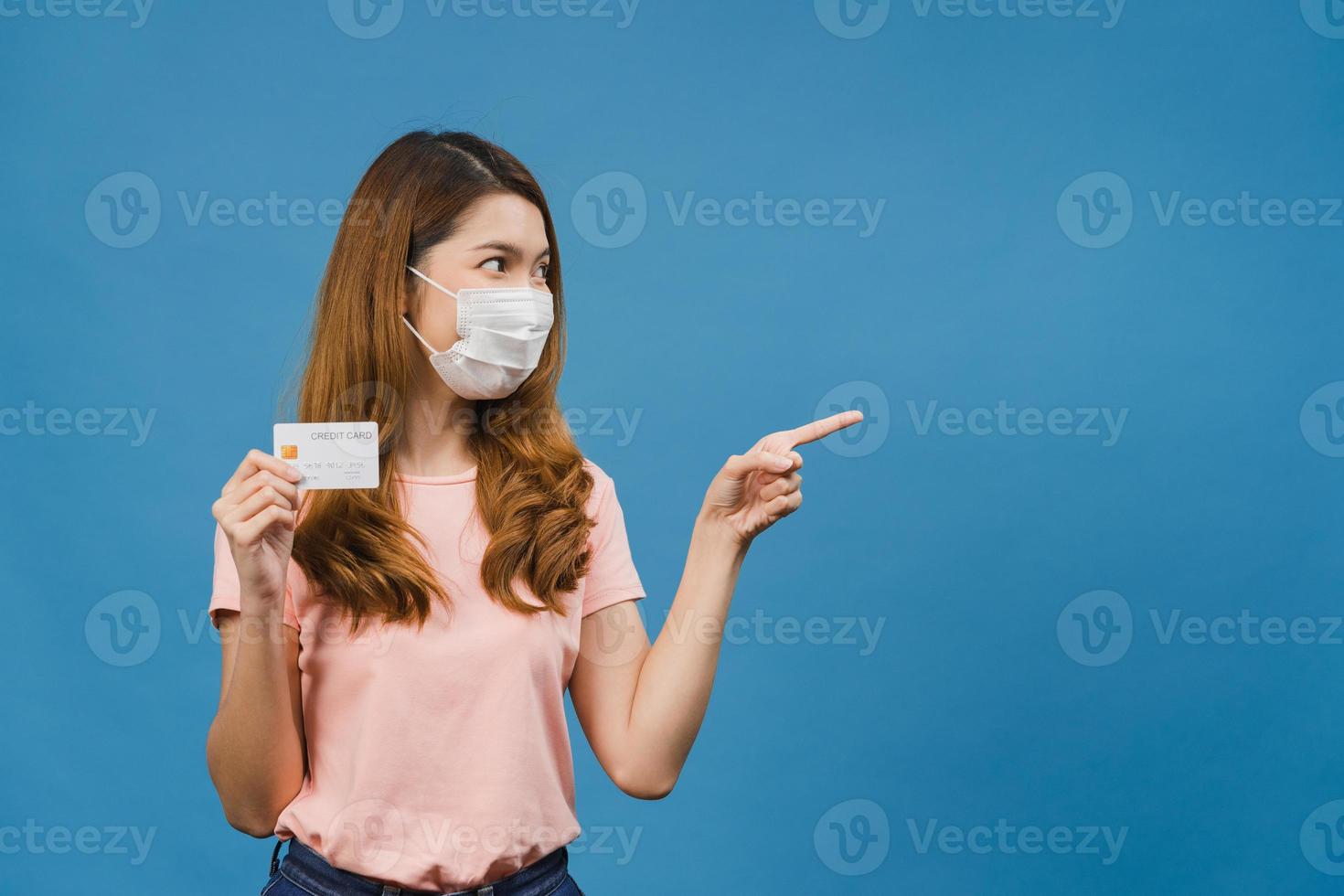 Young Asia lady wearing medical face mask show credit bank card with positive expression, smiles broadly, dressed in casual clothing feeling happiness and stand isolated on blue background. photo