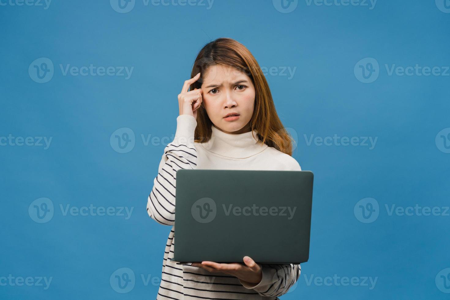 Jovencita asiática usando laptop con expresión negativa, gritando emocionado, llorando emocionalmente enojado en ropa casual y parado aislado sobre fondo azul con espacio de copia en blanco. concepto de expresión facial. foto