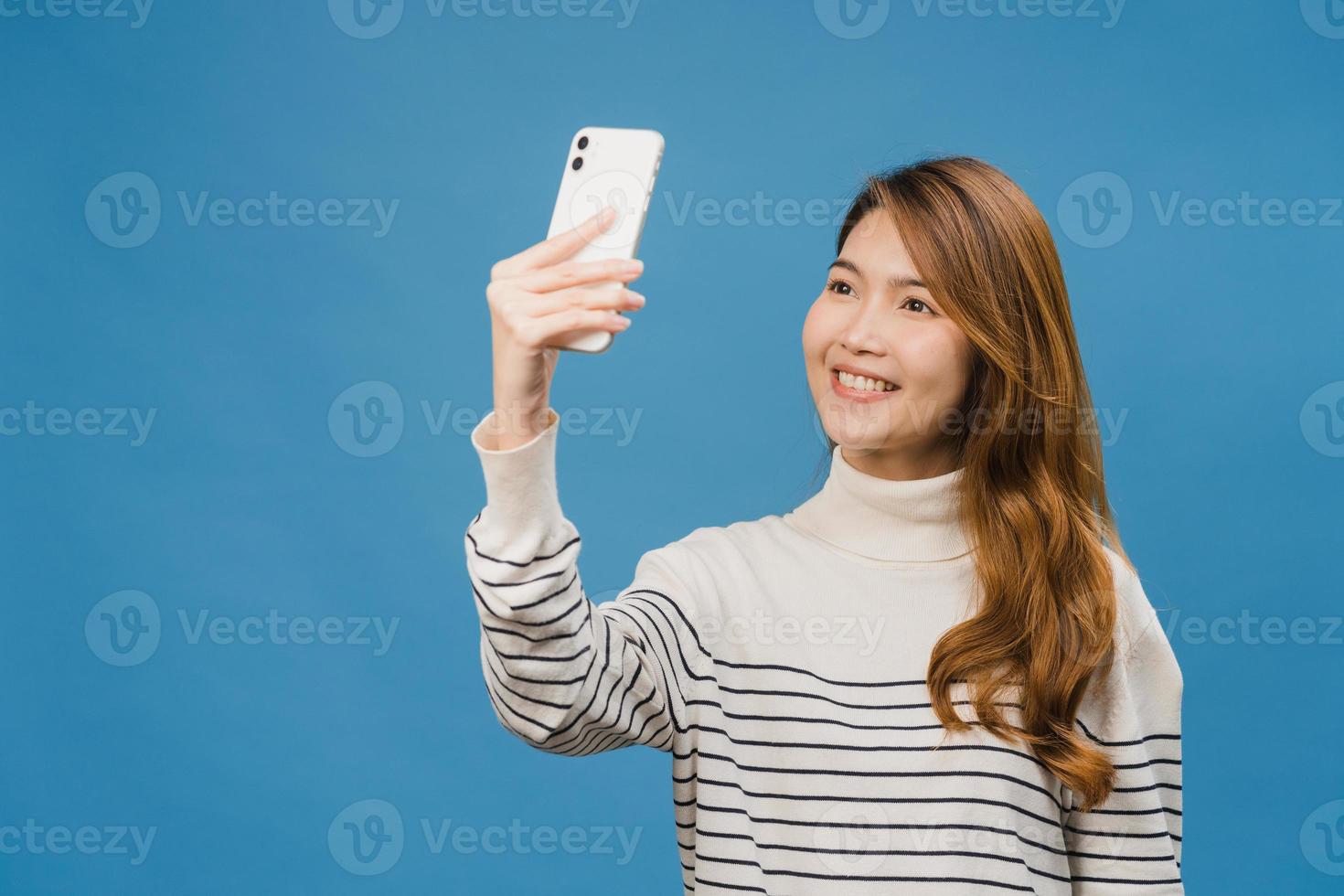 Smiling adorable Asian female making selfie photo on smart phone with positive expression in casual clothing and stand isolated on blue background. Happy adorable glad woman rejoices success.