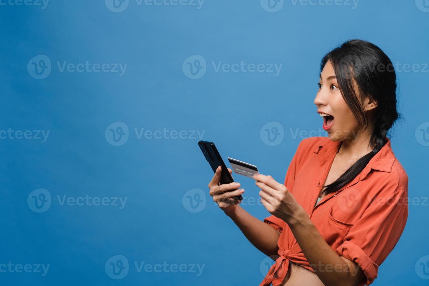 Young Asia lady using phone and credit bank card with positive expression, smiles broadly, dressed in casual clothing and stand isolated on blue background. Happy adorable glad woman rejoices success. photo