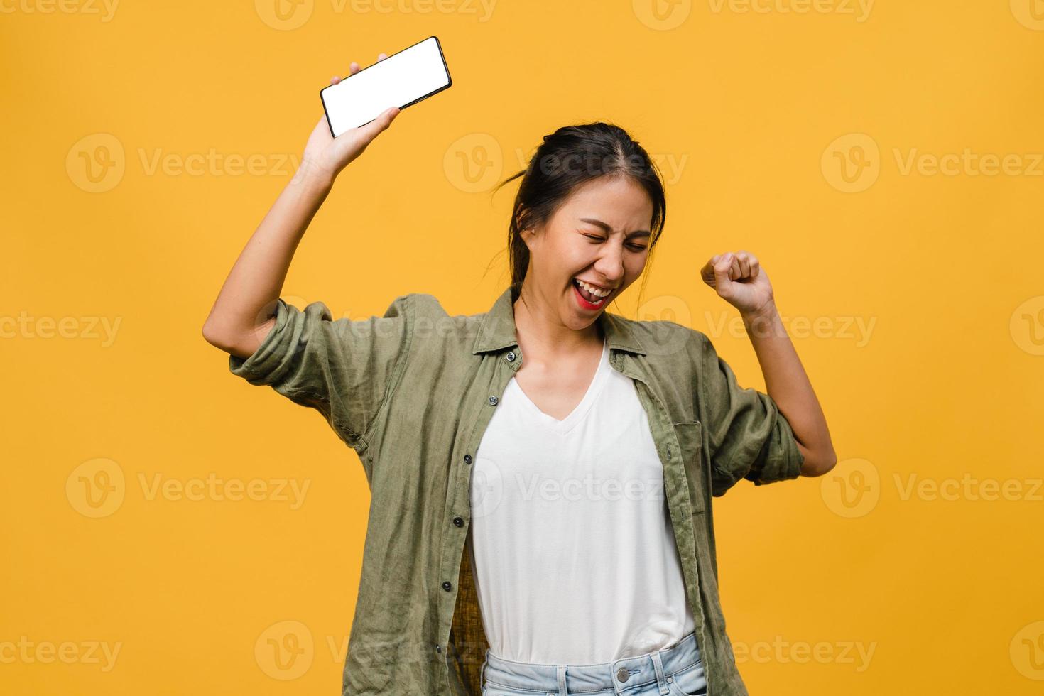 Young Asia lady show empty smartphone screen with positive expression, smiles broadly, dressed in casual clothing feeling happiness on yellow background. Mobile phone with white screen in female hand. photo