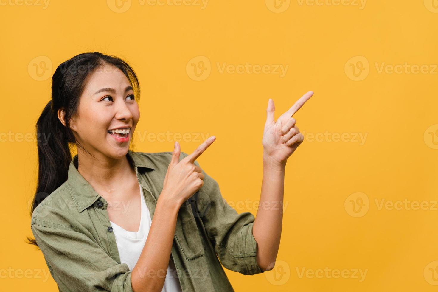 Portrait of young Asian lady smiling with cheerful expression, shows something amazing at blank space in casual clothing and standing isolated over yellow background. Facial expression concept. photo