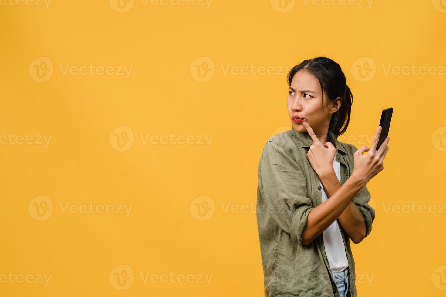 Thinking dreaming young Asia lady using phone with positive expression, dressed in casual cloth feeling happiness and stand isolated on yellow background. Happy adorable glad woman rejoices success. photo