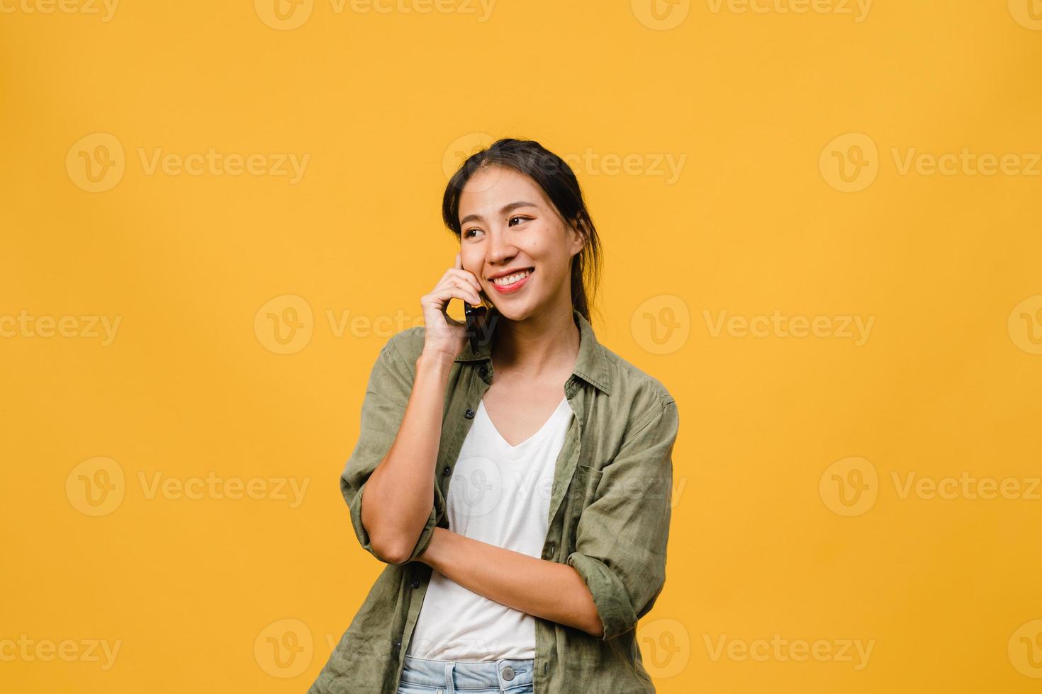 Young Asia lady talk by phone with positive expression, smile broadly, dressed in casual clothing feeling happiness and stand isolated on yellow background. Happy adorable glad woman rejoices success. photo