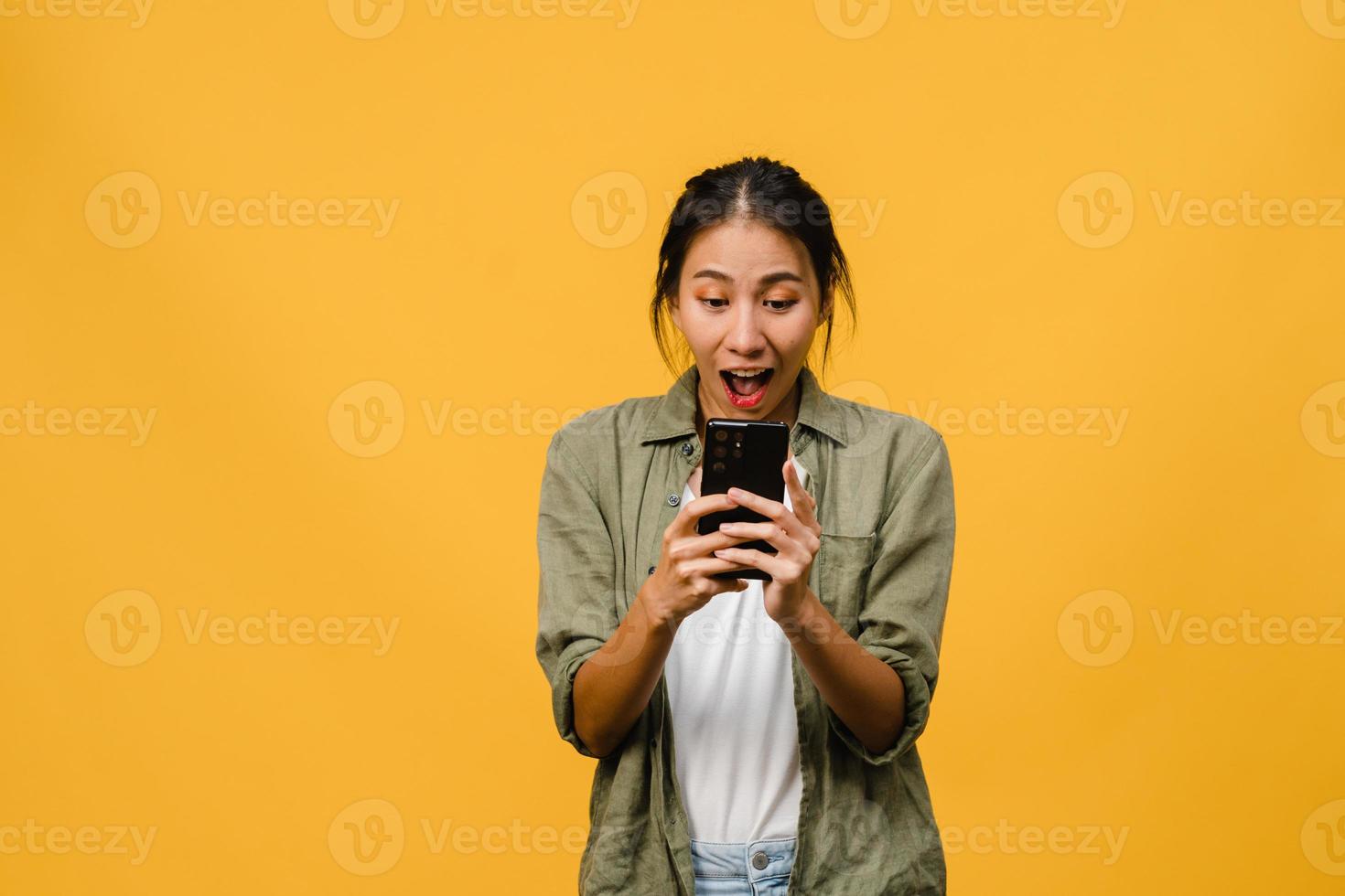 Surprised young Asia lady using mobile phone with positive expression, smiles broadly, dressed in casual clothing and stand isolated on yellow background. Happy adorable glad woman rejoices success. photo