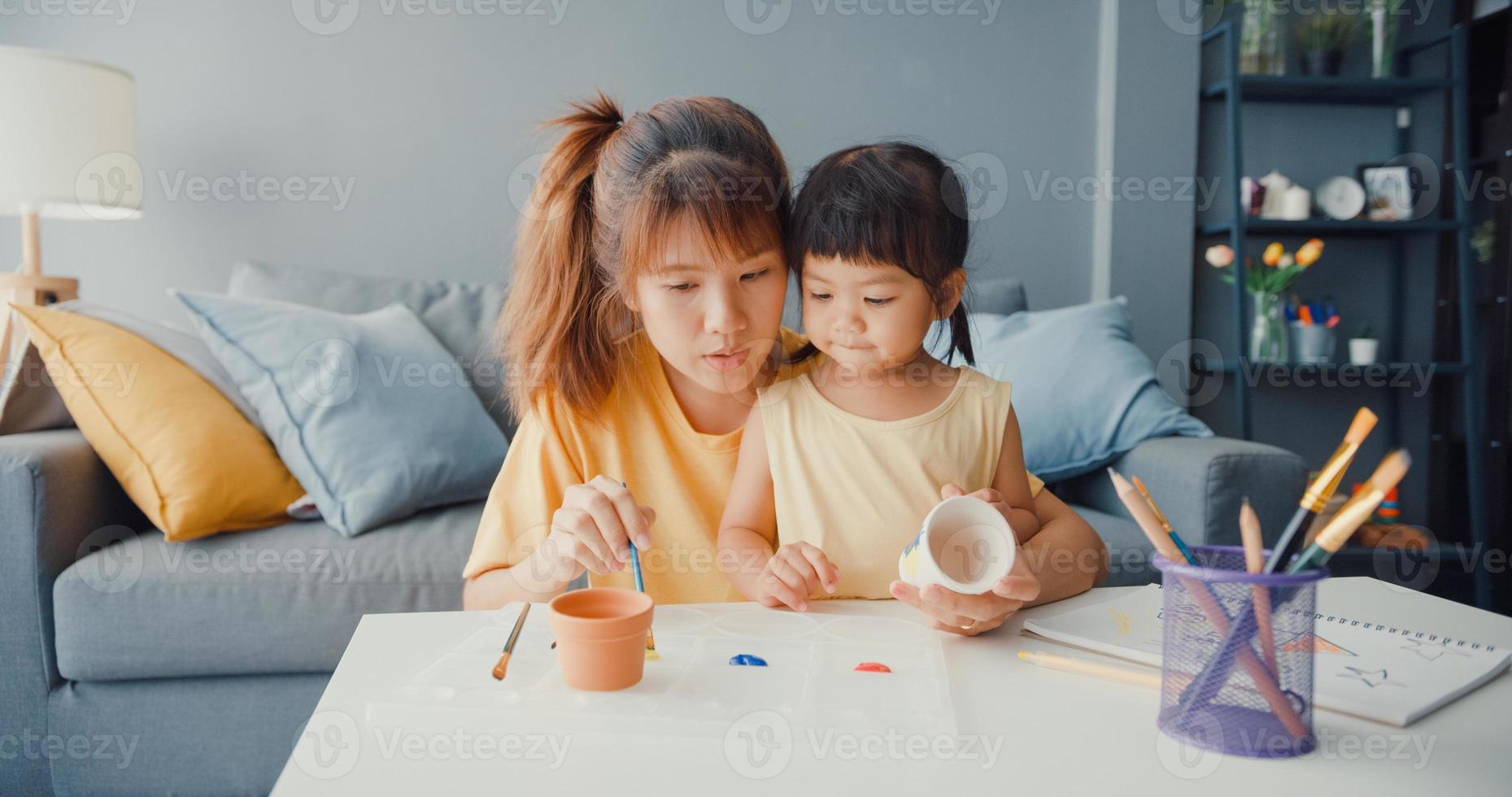 Happy cheerful Asia family mom teach toddler girl paint ceramic pot having fun relax on table in living room at house. Spending time together, Social distance, Quarantine for coronavirus prevention. photo