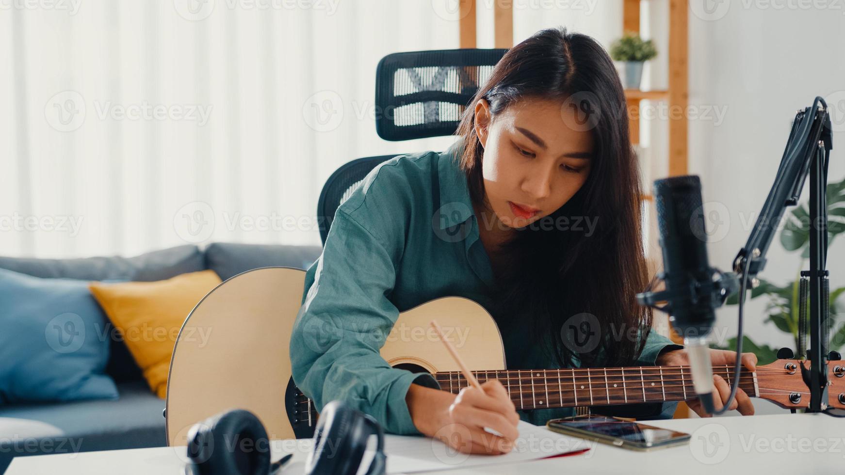 Happy asia woman songwriter play acoustic guitar listen song from smartphone think and write notes lyrics song in paper sit in living room at home studio. Music production at home concept. photo