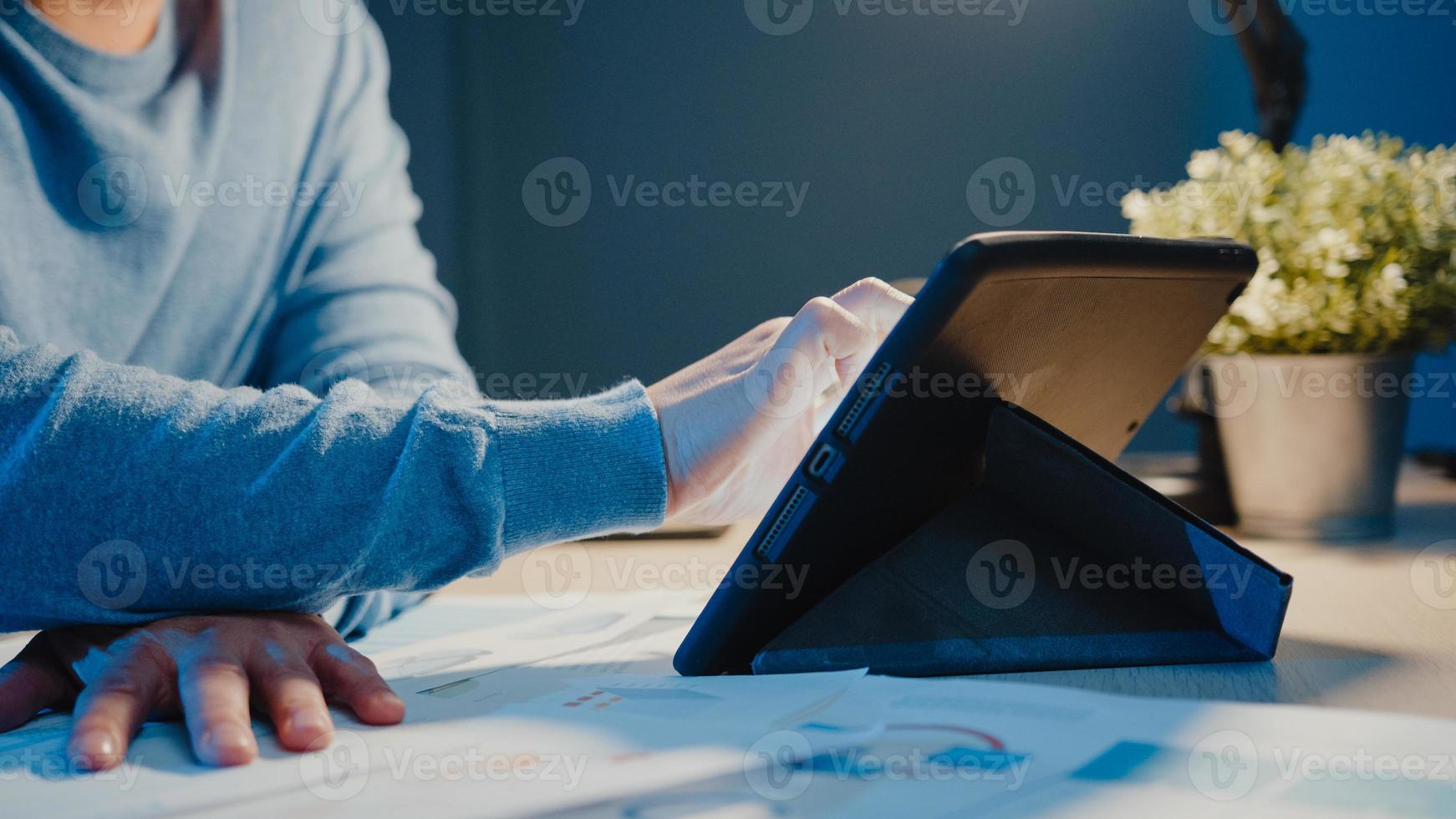 Close-up Asia empresario autónomo trabajo de enfoque escribir en la tableta ocupado con papeleo lleno de gráficos en la sala de estar en casa horas extras por la noche, trabajar desde casa durante el concepto de pandemia de coronavirus. foto